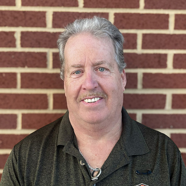 Lee Schantz, Regional Operations Specialist at Arrow Senior Living, smiling in a professional headshot. He has short gray hair, a mustache, and wears a dark collared shirt with a silver chain necklace, standing in front of a red brick wall background.