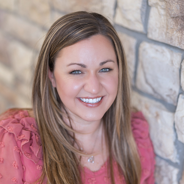 Stacey Russell, Regional Executive Director at Arrow Senior Living, smiling in a professional headshot taken at The Princeton Senior Living community. She has long light brown hair, wears a pink textured blouse, and is posed against a tan stone wall background.