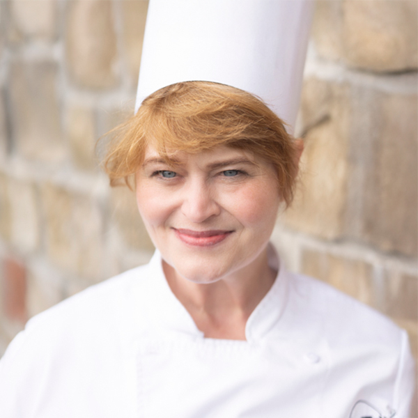 Trisha Ton, Regional Culinary Coordinator at at Arrow Senior Living, wearing a chef's hat and white coat, smiling in front of a stone wall background.