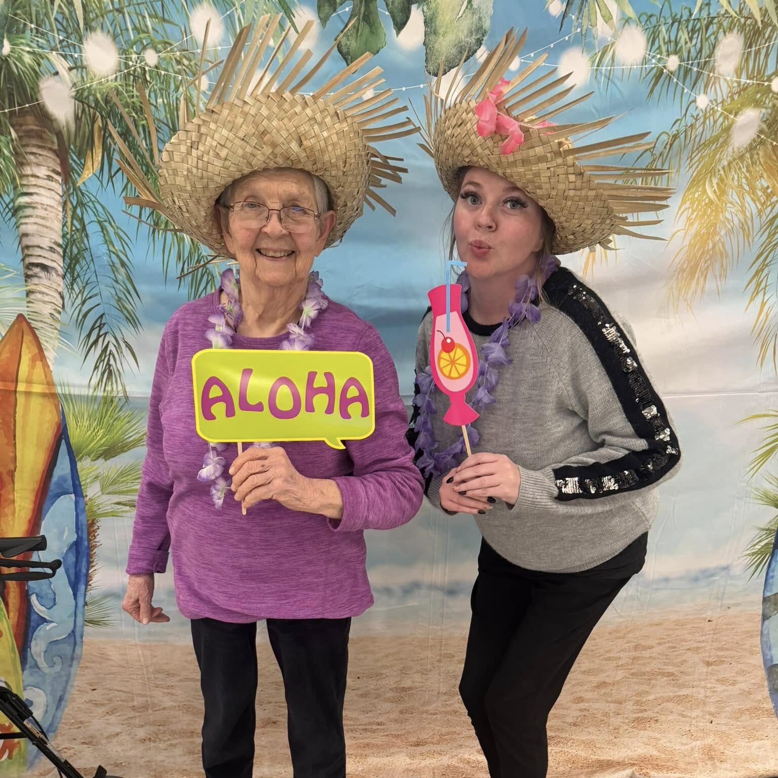 A senior resident and a team member pose in tropical hats at a beach-themed event, holding colorful props while standing in front of a backdrop with palm trees and sand.