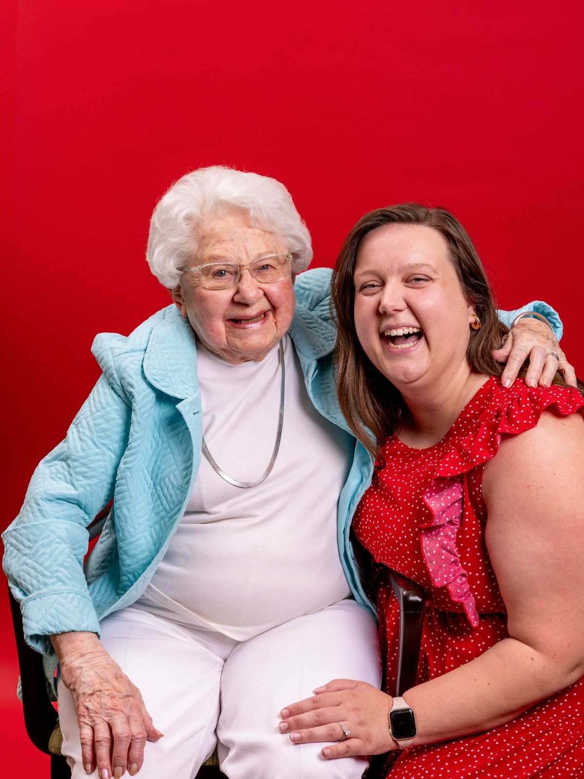 A senior resident in a light blue jacket smiles beside a younger woman in a red dress, both laughing and sitting closely together against a bright red backdrop.