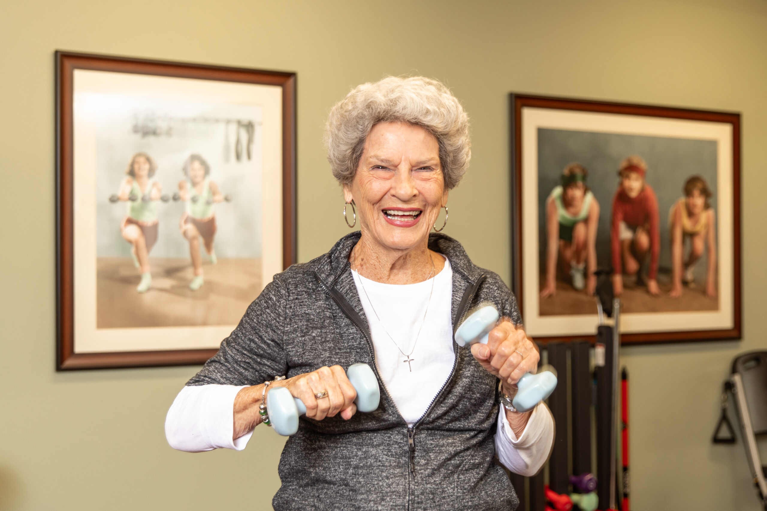 A smiling senior resident lifting small dumbbells in a fitness room, staying active and healthy with exercise as part of a wellness program.