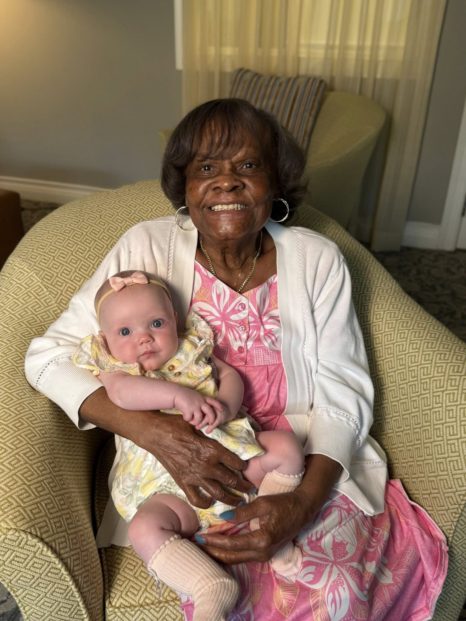 A senior resident smiling while holding a baby on her lap in a cozy chair, capturing a sweet intergenerational moment filled with love and warmth.