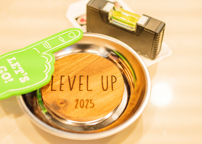 Close-up of a small silver dish holding a round wooden token engraved with “Level Up 2025,” surrounded by a green foam finger reading “Let’s Go!” and a black and yellow leveling tool.