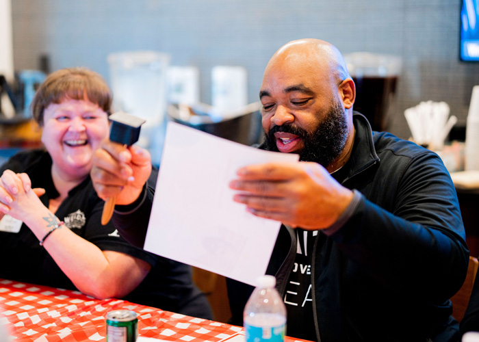 A man laughing as he reads an award certificate at a red-checkered table, holding a small trophy mallet while a coworker beside him smiles during a culinary team celebration.