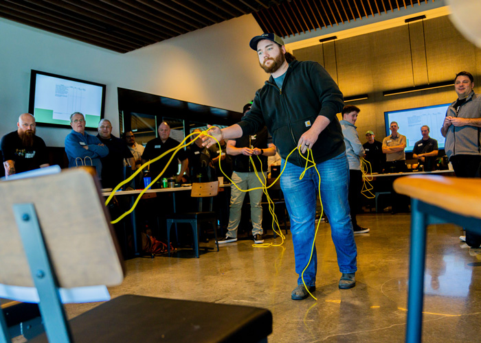 A man in a black hoodie and cap attempts to lasso a chair with bright yellow cords during a team-building challenge, while coworkers watch and laugh in a modern meeting room.