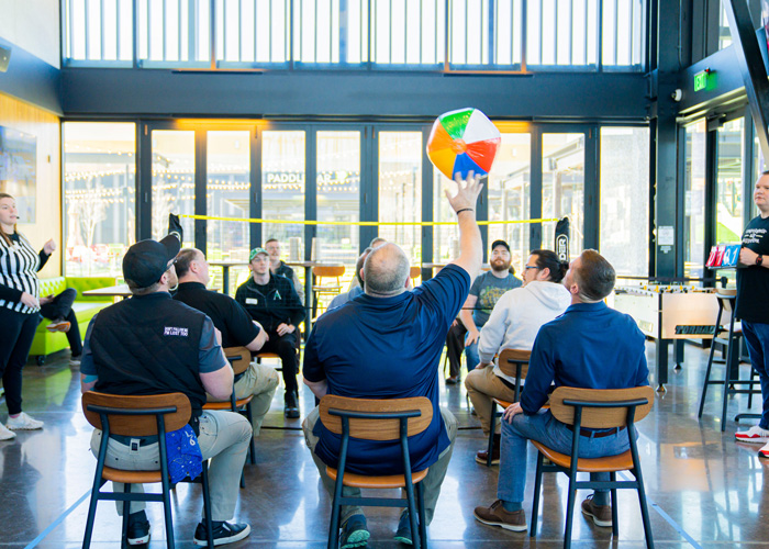 Team members sit in a circle of chairs playing a lively beachball volleyball game indoors, as one player reaches up to hit a colorful ball while others watch and cheer.