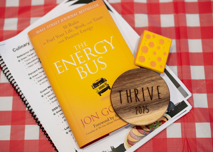 A bright yellow copy of The Energy Bus rests on a red checkered table with culinary training papers, a wooden Thrive 2025 token, and a small yellow polka-dot stress block.