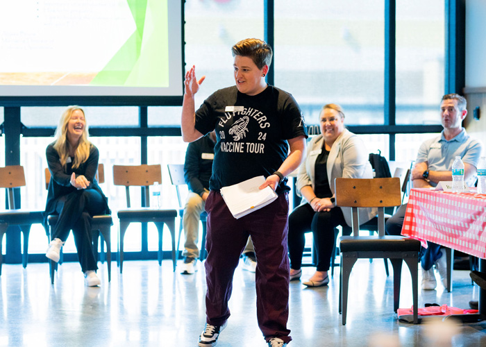 A presenter in a "Flu" Fighters T-shirt speaks confidently while holding papers, standing before seated attendees in a bright room with large windows and a presentation screen.