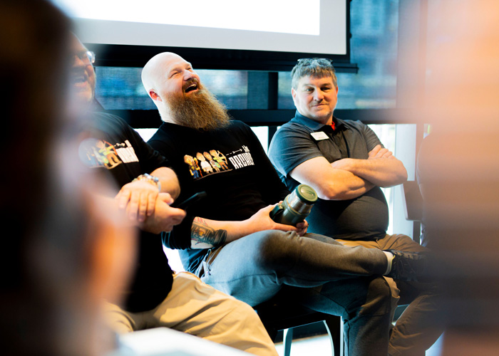 Two team members sit on stools during a Plant Operations session, one laughing with his head back while the other smiles with arms crossed, creating a warm, upbeat moment.