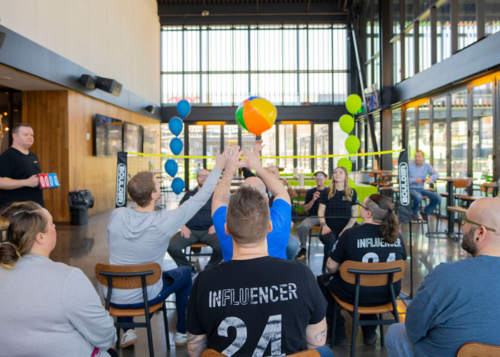 Team members play seated beachball volleyball in a bright open room, reaching up together to hit a colorful ball over a small net decorated with balloons.