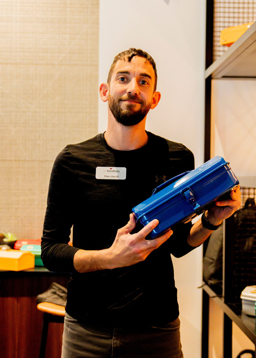A team member at the Plant Operations Summit smiles while holding a bright blue toolbox, standing in a warmly lit room with shelves and supplies in the background.