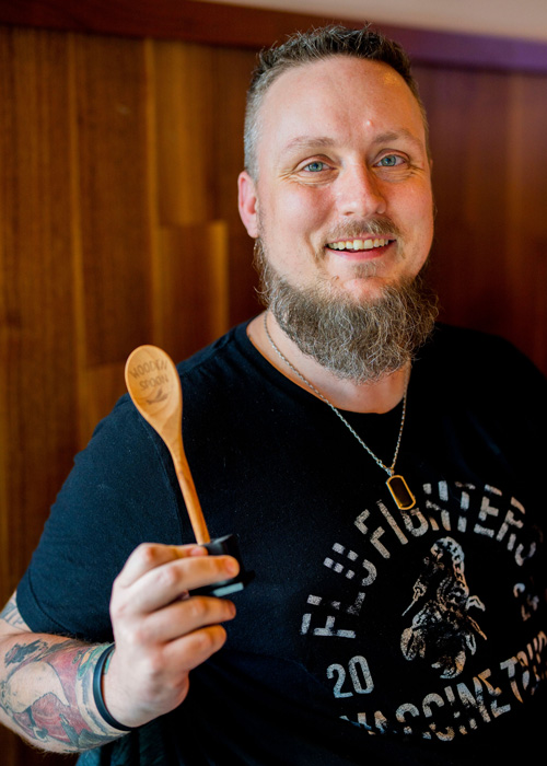 A smiling man with a beard and tattooed arms holds up a wooden spoon award while standing against a warm wood-paneled wall, wearing a black "Flu" Fighters T-shirt.