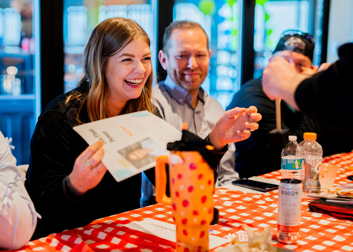 A woman smiles brightly at a red-checkered table as she receives an award certificate, surrounded by coworkers and drinks during a lively culinary team gathering.