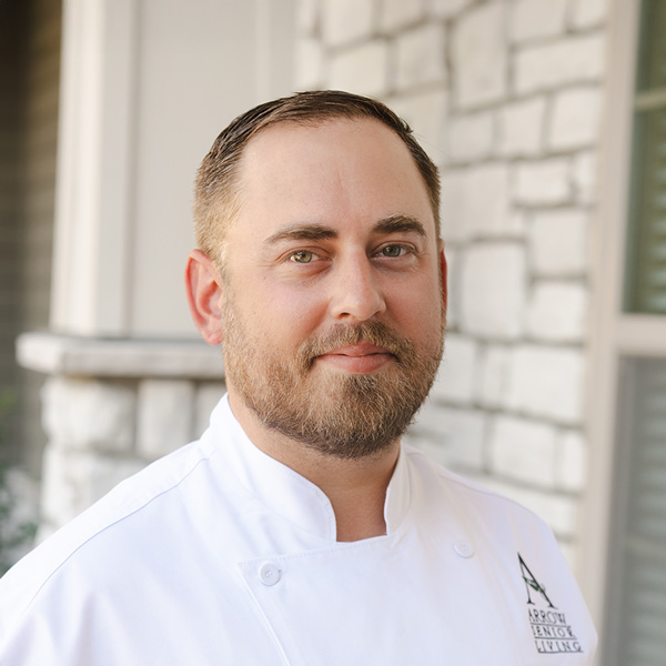 Nick Stromack, Regional Culinary Coordinator at Arrow Senior Living, standing outdoors in a white chef coat, smiling slightly with a stone building in the background.