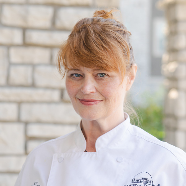 Trisha Ton, Regional Culinary Coordinator at Arrow Senior Living, smiling outdoors in a white chef coat with light stone columns and greenery softly blurred behind her.