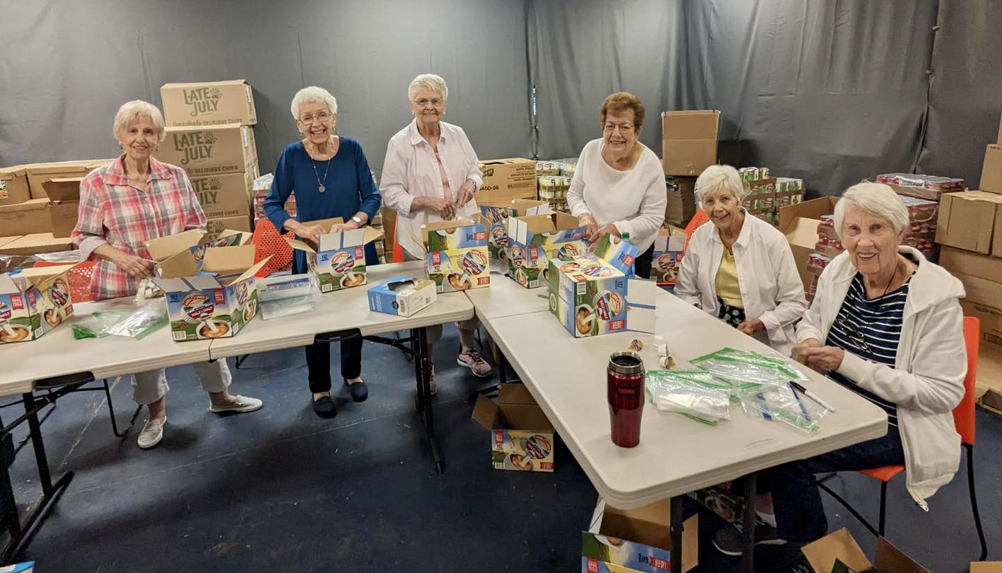 A group of senior residents assembling care packages together, volunteering joyfully at tables surrounded by boxes in a community service project.