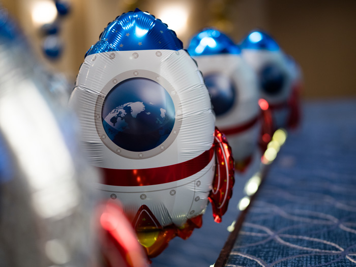 A close-up of metallic rocket-shaped balloons featuring a blue and white design with a globe illustration in the window, arranged in a row on a carpeted surface at an indoor event.