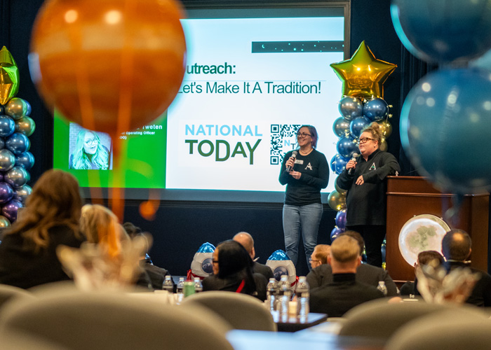 Two speakers present on stage at the 2025 Arrow Summit, surrounded by balloon decorations and an audience of team members focused on the National Today slide.