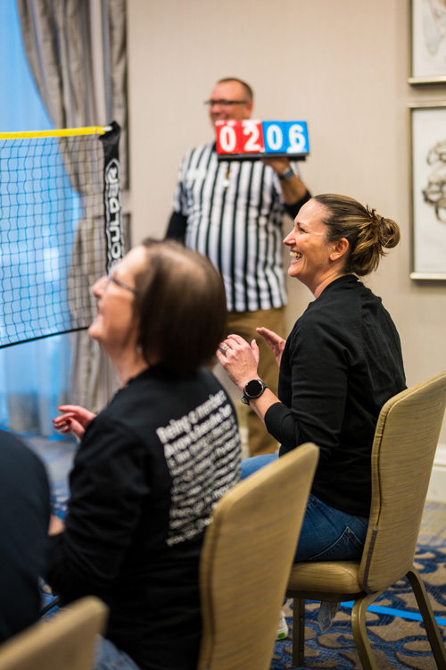 Team members laugh during a friendly indoor chair volleyball match at the 2025 Arrow Summit, as a referee holds up the game score in the background.