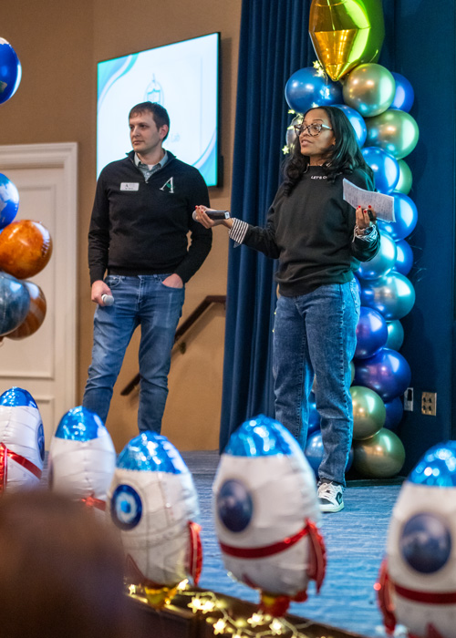 Two Arrow Senior Living team members present on stage at the 2025 Arrow Summit, surrounded by colorful balloon rockets and a space-themed Mission Control backdrop.