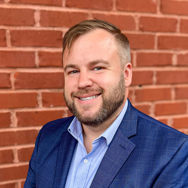 Adam Kerst, Financial Planning and Analysis team member at Arrow Senior Living Home Office, smiling in a blue blazer while standing in front of a brick wall background.