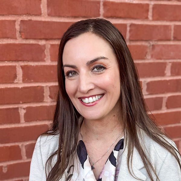Lauren Horn, Managing Director of Wellness Integration at Arrow Senior Living Home Office, smiling in a professional headshot, wearing a light blazer and patterned blouse, standing against a red brick wall.