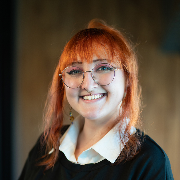 Ashleigh Koenig, Arrow Memory Care Director at Arrow Senior Living Home Office, smiling in a professional headshot with glasses, red hair, and a softly lit indoor background.