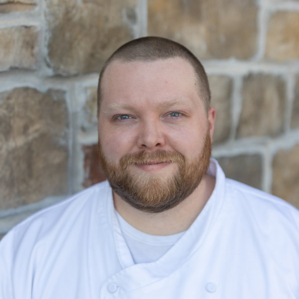 Sean Jensen, Regional Culinary Coordinator at Arrow Senior Living, in a professional headshot wearing a white chef coat, standing against a stone wall with a calm, confident expression.