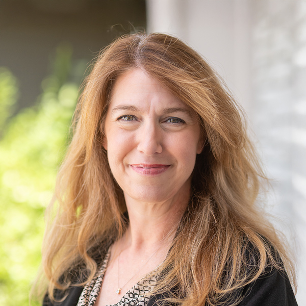 Amy Byergo, Regional Executive Director at Arrow Senior Living, smiling with long light brown hair, wearing a black blazer, standing outdoors near greenery.