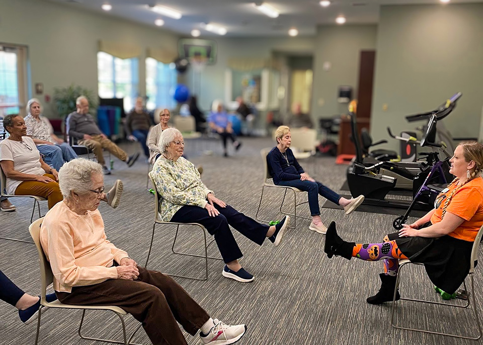 Senior residents seated in the fitness center participate in a fitness session led by a team member.