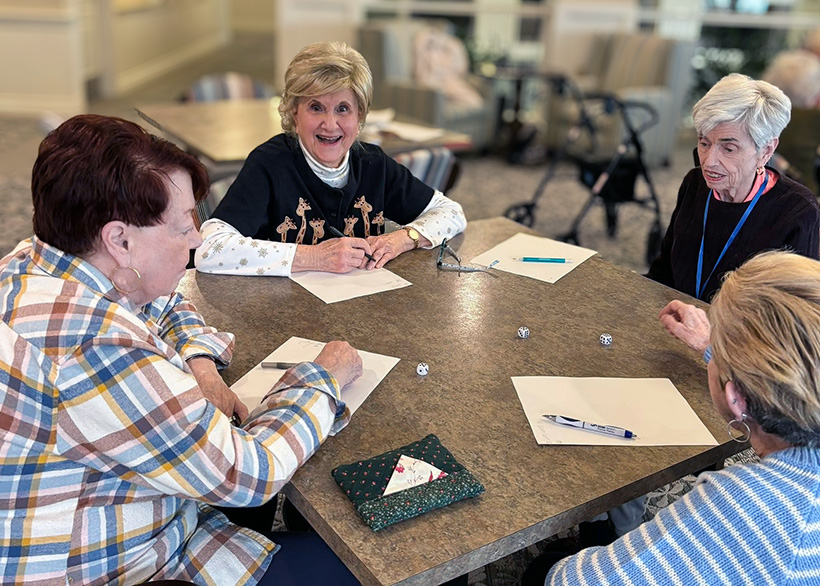 Senior women gathered around a community table in a warm, inviting common space enjoy a tabletop game together.