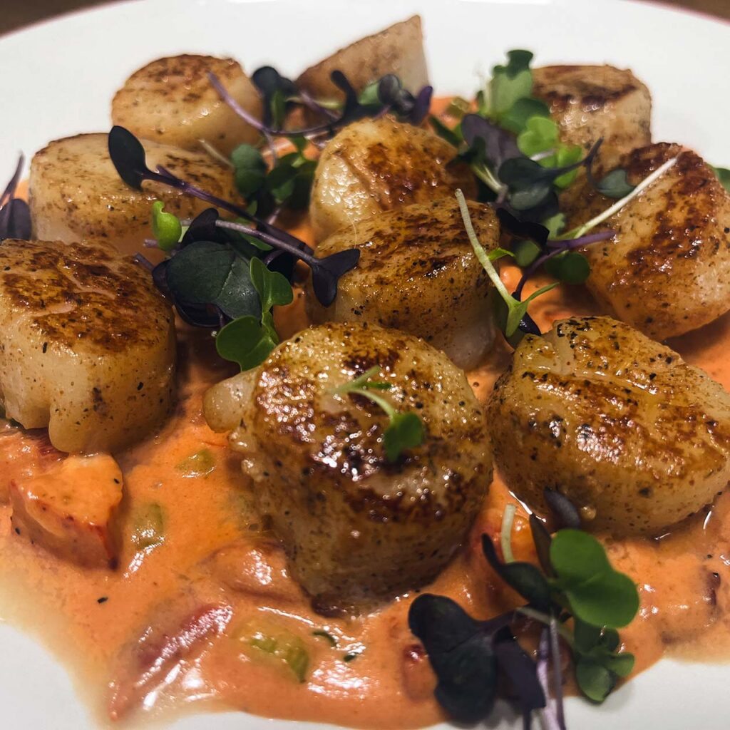 A chef-prepared dish of scallops seared to golden brown, resting on a rich sauce with fresh vegetables and topped with micro-greens.