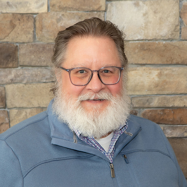 Charlie Johnson, Creative Project Coordinator at Arrow Senior Living, smiling in a professional headshot. He wears glasses and a blue quarter-zip pullover over a patterned collared shirt, with a stone wall background.”