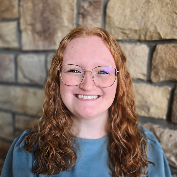 Jordyan Craig, Director of Community Payroll at Arrow Senior Living Home Office, smiling in front of a stone wall, wearing glasses and a blue top.