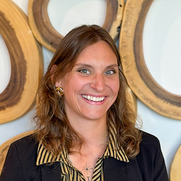 Kelly Voss, Arrow Senior Living Director for Home Office, smiling in front of decorative wooden wall art, wearing a black blazer and striped blouse.