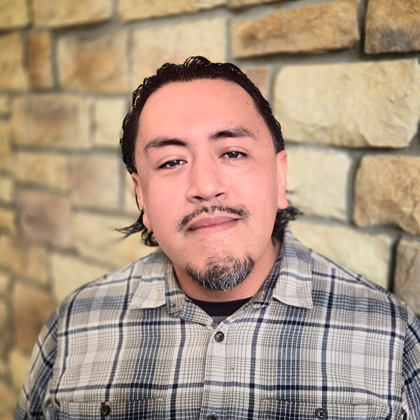 Ernesto Perez, Plant Operations Director on the Arrow travel team at Arrow Senior Living, standing in front of a stone wall, wearing a gray plaid button-down shirt.