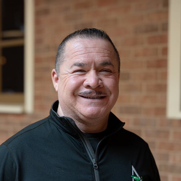 Humberto Barraza, Regional Director of Culinary Compliance at Arrow Senior Living Home Office, smiling in a professional headshot, wearing a black zip-up jacket, with a softly blurred brick background.