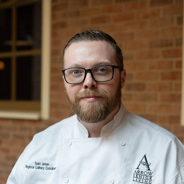 Sean Jensen, Regional Culinary Coordinator at Arrow Senior Living Home Office, smiling in a professional headshot, wearing glasses and a white chef coat, with a softly blurred brick and window background.