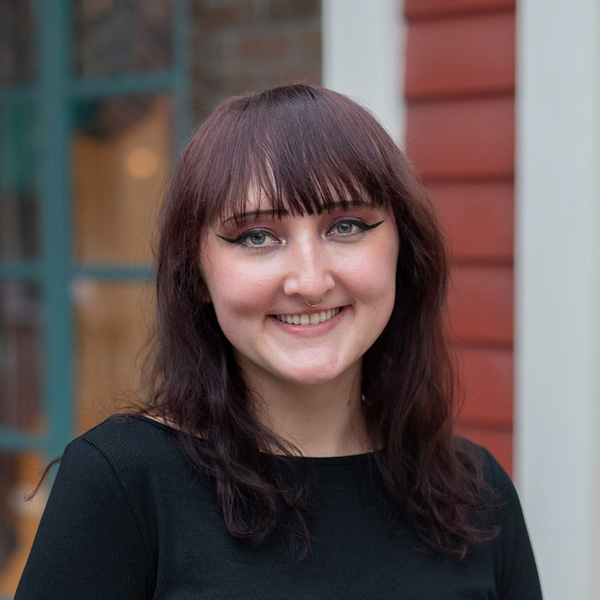 Ashleigh “Ash” Koenig, Arrow Memory Care Director at Arrow Senior Living Home Office, smiling in a professional headshot, wearing a black top, with a softly blurred outdoor background featuring brick and window details.