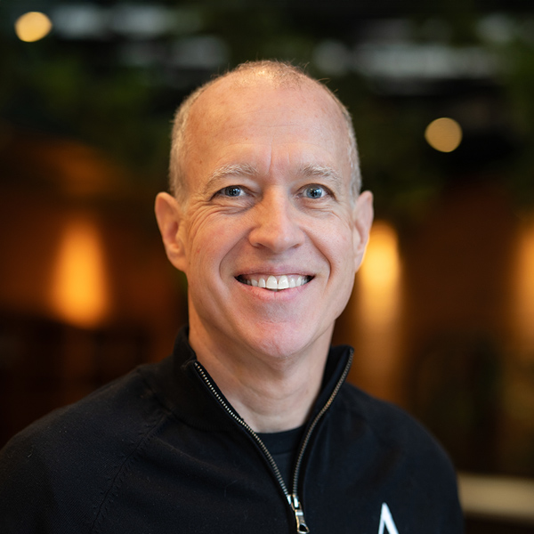 Dan Shields, Regional Executive Director at Arrow Senior Living, smiling in a professional headshot, wearing a black quarter-zip, with a softly blurred indoor background featuring warm lighting and greenery.