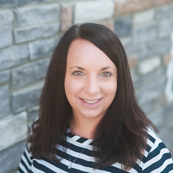 Danielle Kencson, Regional Executive Director at Arrow Senior Living, smiling in a professional headshot, wearing a striped top, with a softly blurred stone wall background.