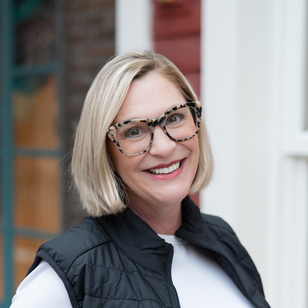 Elizabeth Thompson, Senior Regional Director of Memory Care at Arrow Senior Living, smiling in a professional headshot, wearing glasses and a black vest, with a softly blurred outdoor background featuring brick and window details.