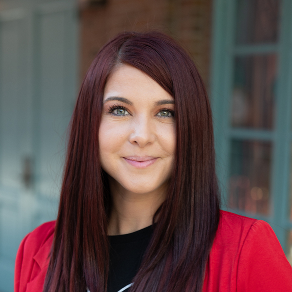 Ellen Cavazos, Regional Wellness Coordinator at Arrow Senior Living, in a professional outdoor headshot with long auburn hair, bright eyes, a red blazer, and softly blurred brick and teal window details in the background.