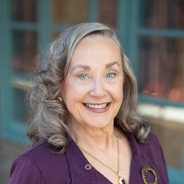Erin Winstead, Executive Director community support for Arrow Senior Living Home Office, smiling in a professional outdoor headshot with curled gray hair, bright blue eyes, a purple top, and softly blurred teal window details in the background.