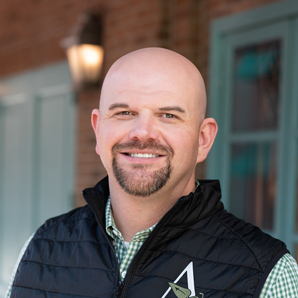 Justin Ladd, Portfolio Manager at Arrow Senior Living Home Office, smiling in a professional outdoor headshot with a bald head, goatee, black vest, and softly blurred brick and teal window details in the background.