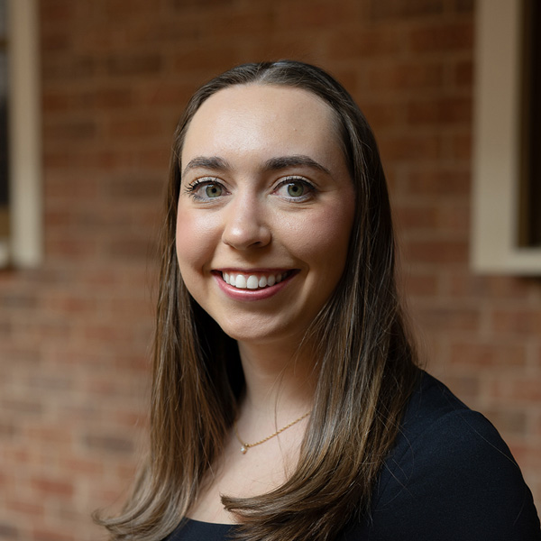Karli Arganese, Wellness Analyst at Arrow Senior Living Home Office, smiling in a professional headshot, wearing a black top, with a softly blurred brick and window background.