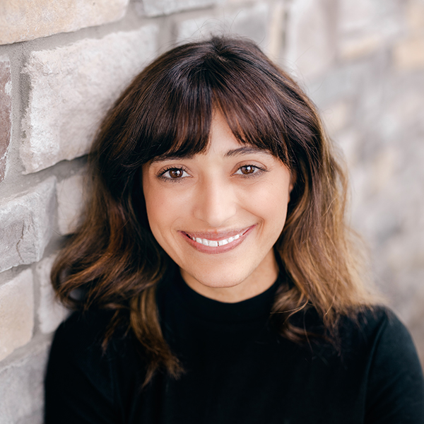 Leah Gallas, Arrow Resident Services Director at Arrow Senior Living Home Office, smiling in a professional headshot with dark shoulder-length hair and bangs, wearing a black top against a light stone wall background.