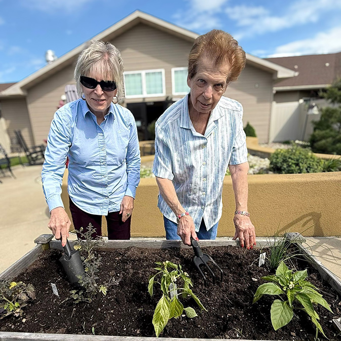 A man and woman tend a raised bed of plants on a sunny day, gardening tools scooping up enriched potting soil.