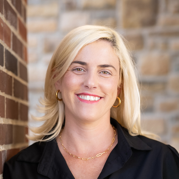 Michelle Graham, Arrow Wellness Director at Arrow Senior Living Home Office, smiling in a professional headshot with blonde hair, gold hoop earrings, and a softly blurred brick and stone background.